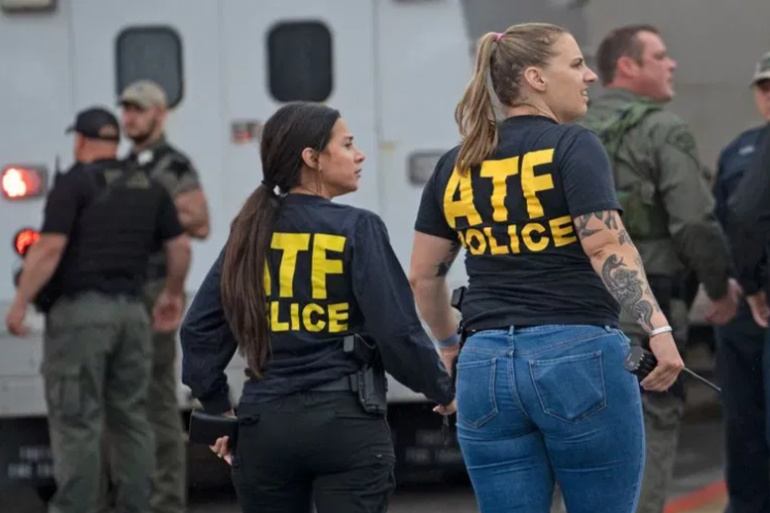 Two ATF police officers stand together while other officers are in the background near an emergency vehicle.