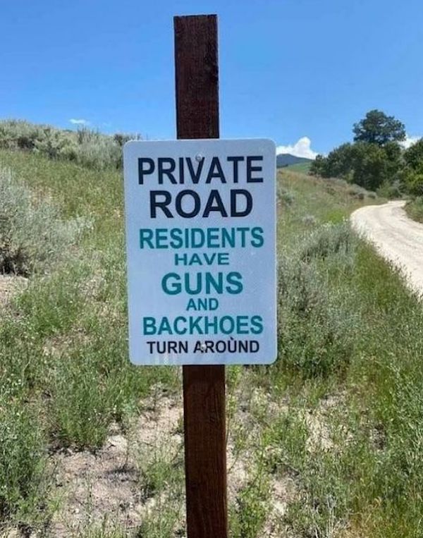 A sign on a post reads, "Private Road. Residents Have Guns and Backhoes. Turn Around." The road and grassy surroundings are visible in the background.