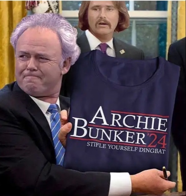 A man with a purple wig holding a shirt that reads "Archie Bunker '24 Stifle Yourself Dingbat" while another man with a mustache stands in the background.