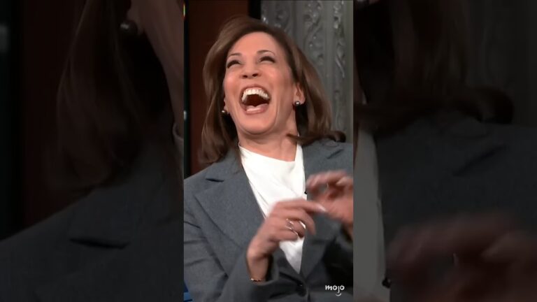 A person wearing a gray blazer and white shirt is laughing heartily with their head tilted back.
