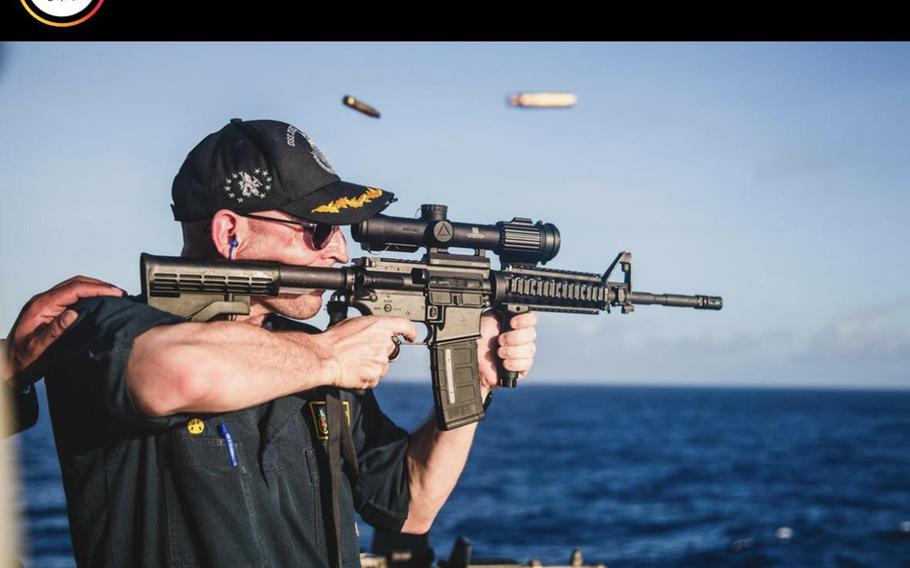 A person in a black cap and ear protection is firing a scoped rifle over open water with spent cartridges ejected in mid-air.