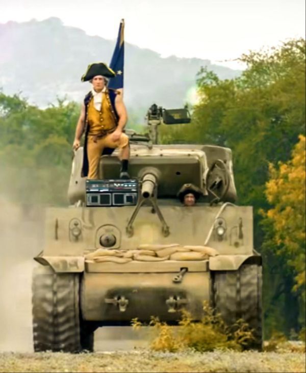 A person in historical military attire stands on a moving tank, with another person inside. The tank is on a dirt road, surrounded by trees.