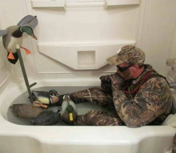 Person in camouflage outfit sitting in a bathtub filled with water, surrounded by duck decoys, and holding a camera with a large lens.