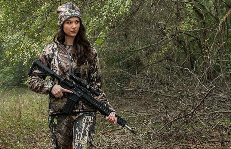 Person in camouflage clothing holding a rifle, walking through a wooded area.