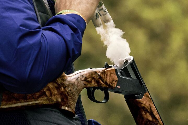 Person wearing a blue jacket holds a smoking shotgun with wood finish, outdoors.