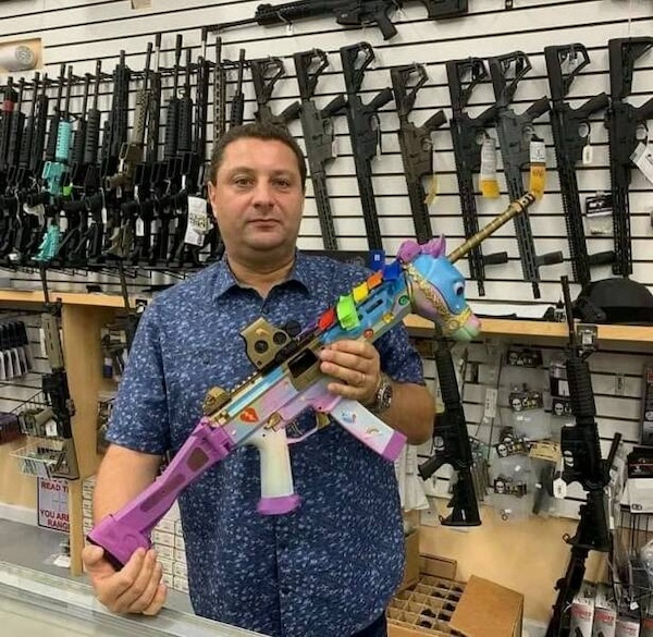 A man stands in a gun store holding a brightly colored, toy-like rifle with a unicorn design; various black rifles hang on the wall behind him.