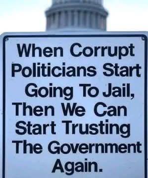 A sign reads, "When corrupt politicians start going to jail, then we can start trusting the government again," with a government building blurred in the background.