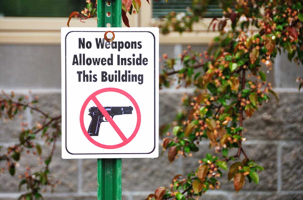 A sign outside a building reads "No Weapons Allowed Inside This Building" with an image of a handgun crossed out.