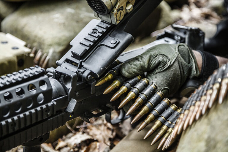 A gloved hand loads a belt of ammunition into a machine gun outdoors, with rocks and leaves visible in the background.