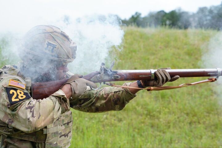 A soldier in camouflage uniform and helmet fires a musket, producing smoke, while standing outdoors on a grassy field.