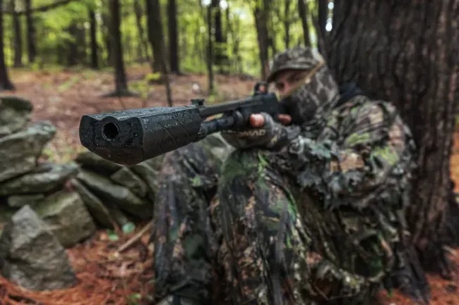 Person in camouflage gear sitting against a tree in a forest, aiming a shotgun fitted with a suppressor.