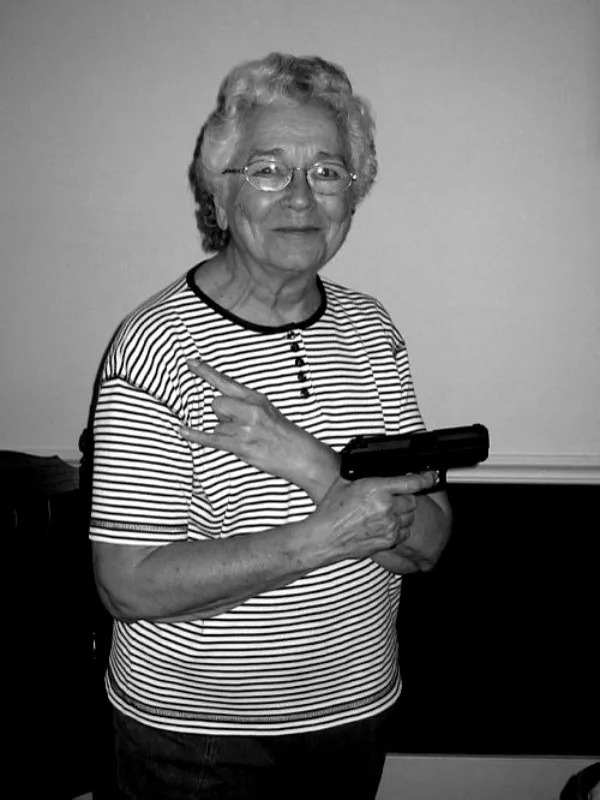 An older woman with glasses, wearing a striped shirt, holds a handgun and makes a peace sign with her other hand. The photo is in black and white.
