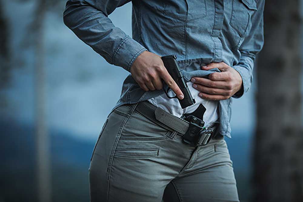 Person drawing a handgun from a holster on their waistband, wearing a denim shirt and gray pants, with an outdoor background.