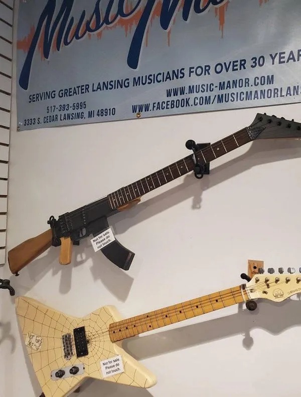 Two electric guitars are mounted on a wall; the top guitar is designed to resemble an assault rifle, and the bottom guitar has a web-like pattern on its body.