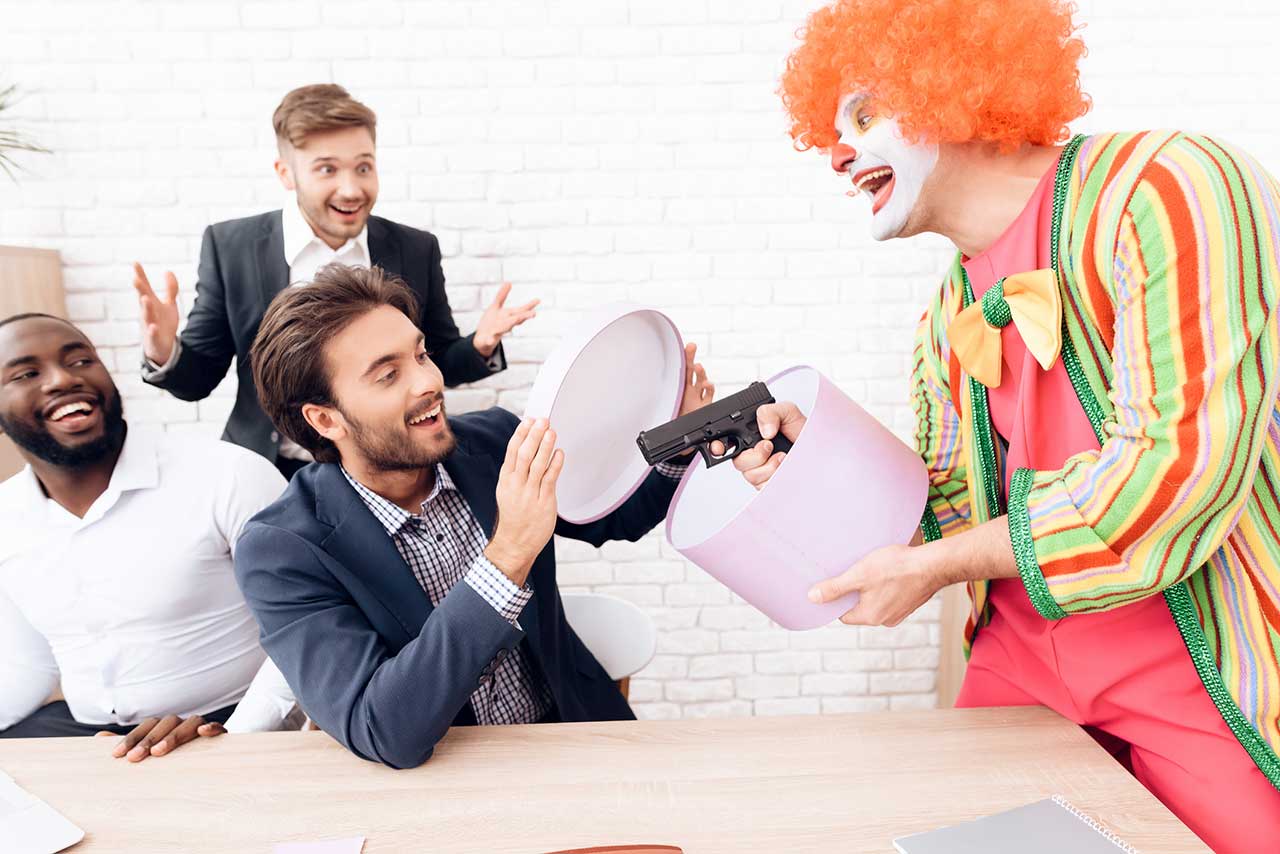 A clown in colorful costume reveals a toy gun from a gift box to surprised men in business attire during an office meeting.