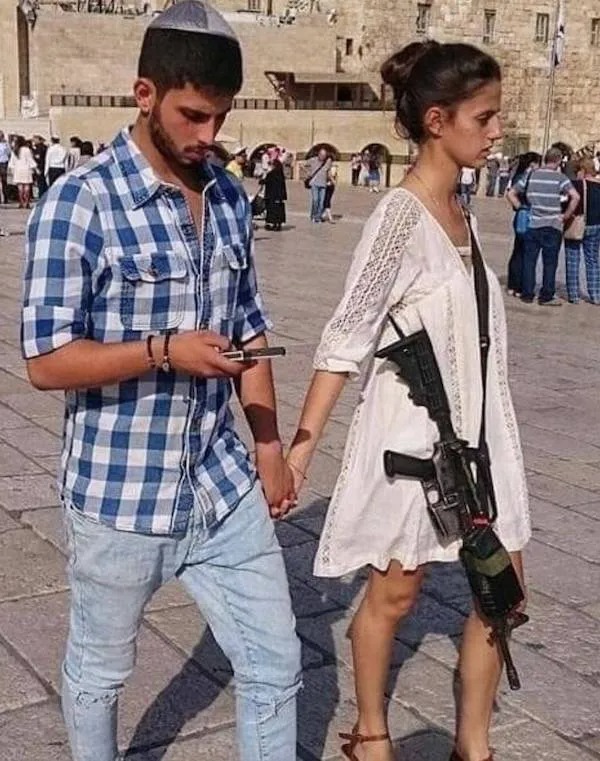 A man and a woman holding hands walk outside; the man looks at his phone while the woman carries a rifle slung over her shoulder.