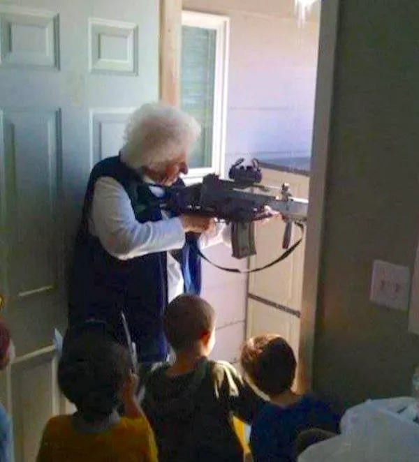 An elderly woman holds a large rifle near a doorway while three children stand nearby and watch.