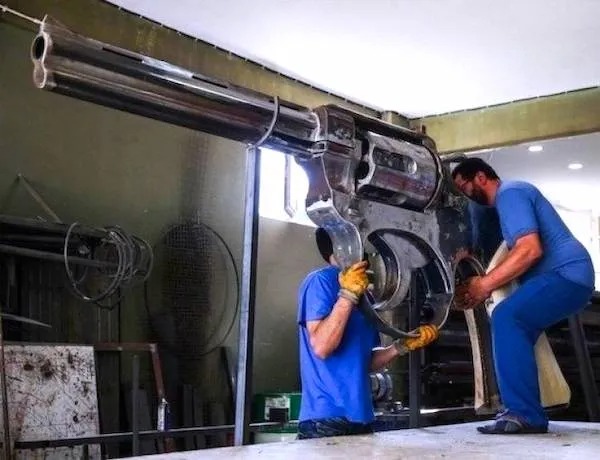 Two workers in blue uniforms assemble a giant metallic revolver replica in an indoor workshop.