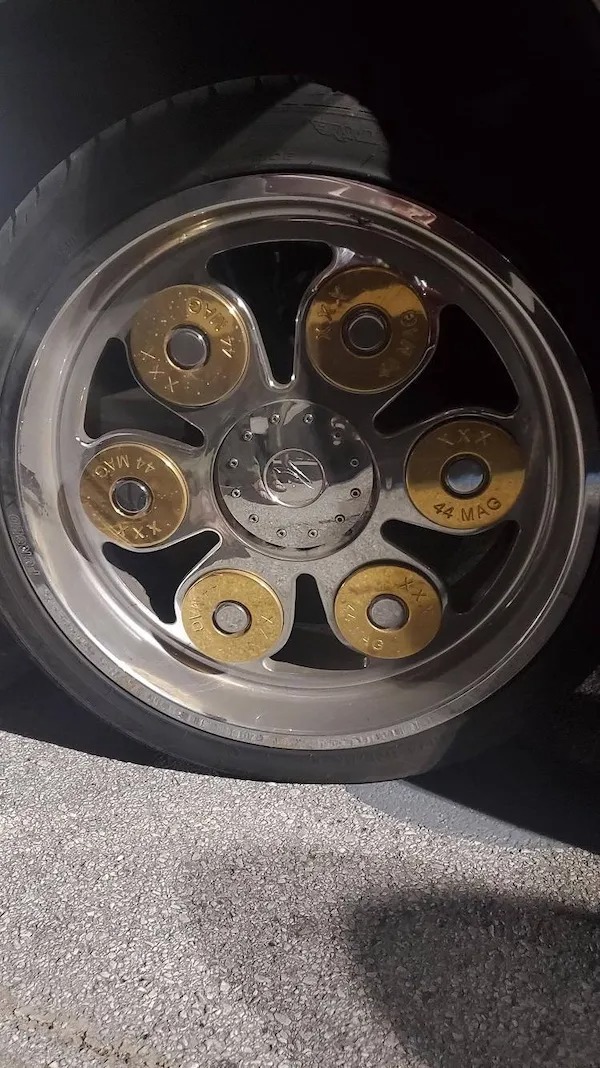 Car wheel rim designed to resemble six large brass .44 Magnum bullet casings, with realistic markings, against an asphalt background.