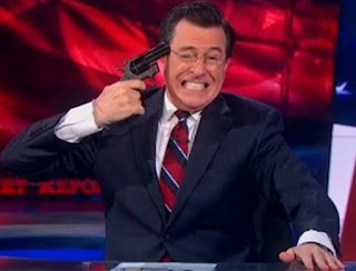A man in a suit and glasses grimaces while holding a fake gun to his head on a television set.