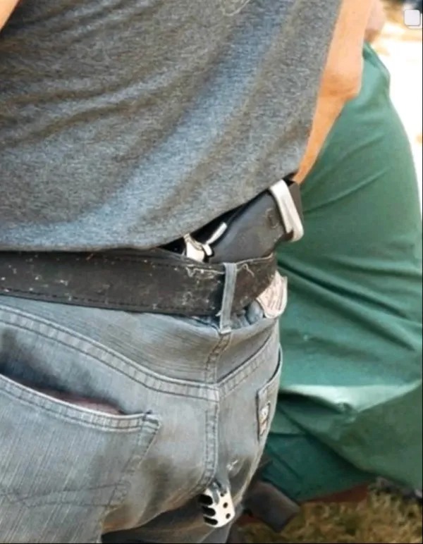 A close-up of a person wearing jeans and a belt with a holstered handgun tucked into the waistband. Another person in green clothing is partially visible in the background.