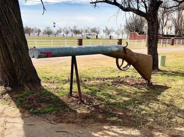 A large sculpture of a shotgun made from metal and wood is mounted between two trees in a grassy outdoor area.