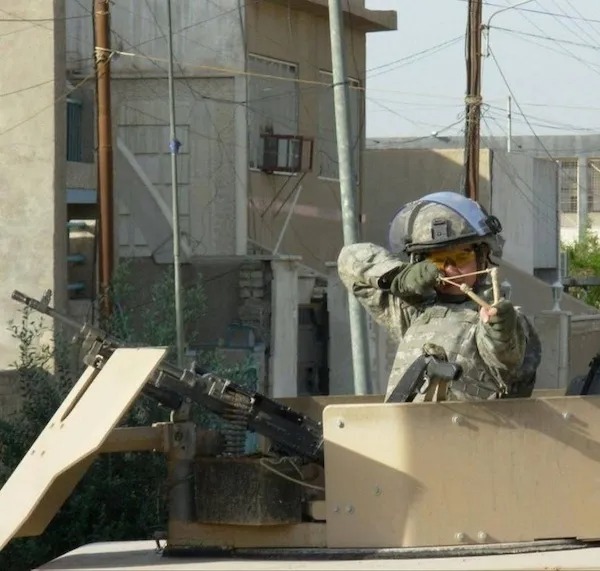 A soldier in uniform inside an armored vehicle aims a slingshot while wearing a helmet and protective gear in an urban setting.