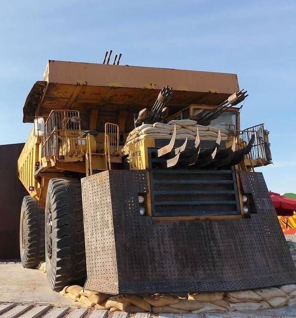 A large, heavily modified dump truck with reinforced armor, sandbags, and multiple mounted weapons on its front, parked outdoors under a clear sky.