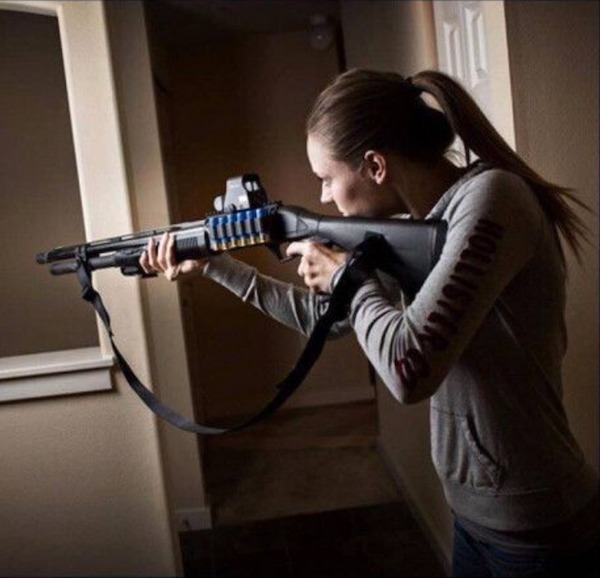 A person in a gray hoodie aims a shotgun with a scope inside a dimly lit hallway.