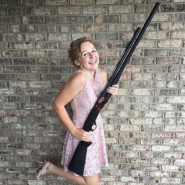 Young woman in a pink lace dress poses smiling against a brick wall, holding a large black shotgun and standing on one foot.