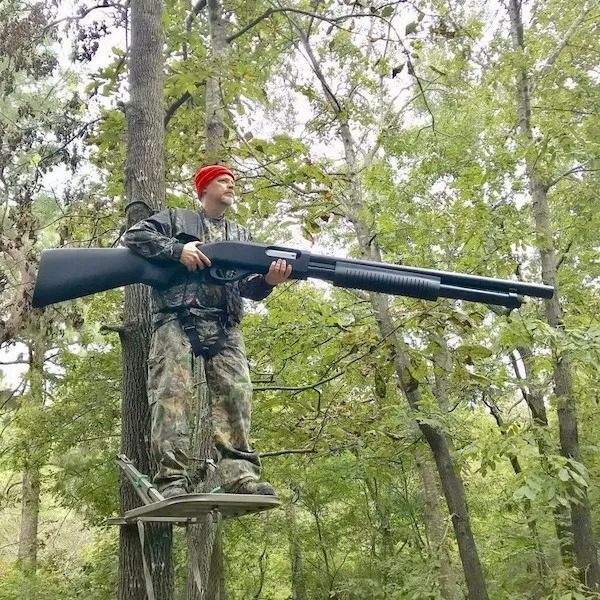 A person in camouflage and a red hat stands on a tree stand in a forest holding an oversized shotgun.