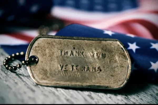 A close-up of a dog tag with "THANK YOU VETERANS" engraved, lying on a surface with an American flag in the background.