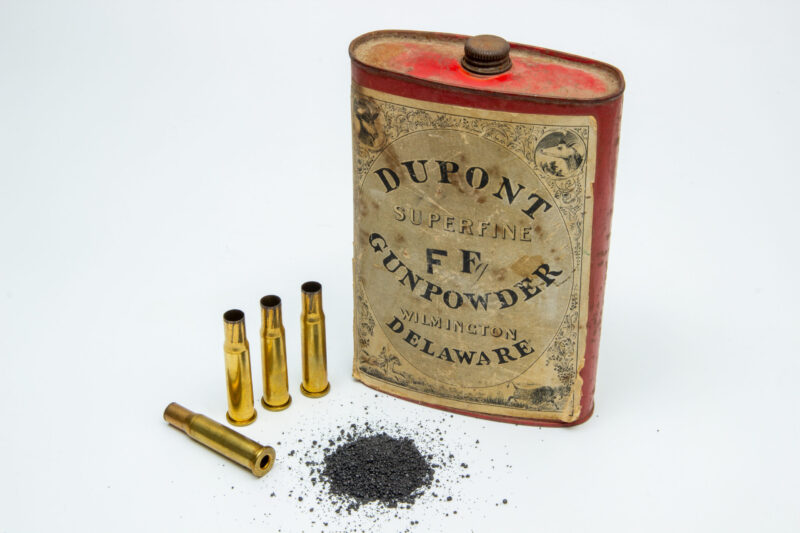 A vintage DuPont gunpowder tin is displayed next to four empty brass bullet casings and a small pile of loose gunpowder on a white background.