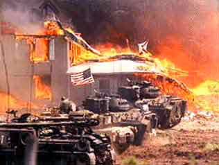 Military tanks in front of a burning building with an American flag, heavy smoke, and flames visible in the background.