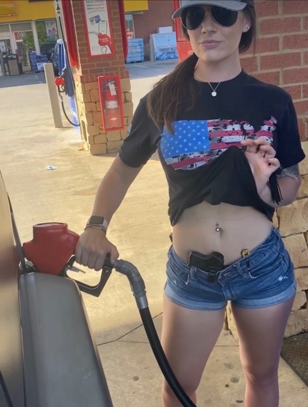 A woman wearing sunglasses and a T-shirt with an American flag design is pumping gas. She lifts her shirt to reveal a handgun in a holster on her waistband.