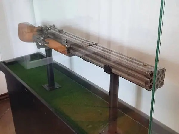 A multi-barreled firearm with a wooden stock displayed horizontally in a glass case on a green surface.