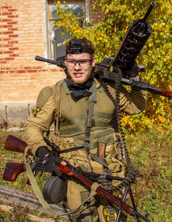 A person in military gear holds two guns and wears protective glasses, a helmet camera, and tactical equipment, standing outdoors near a brick building with autumn foliage in the background.