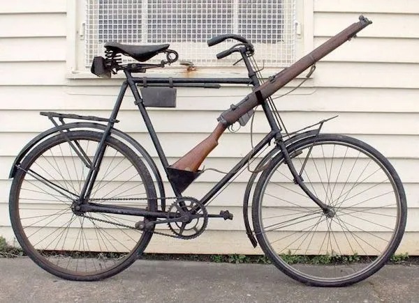 A vintage bicycle with a rifle mounted horizontally above the frame, positioned in front of a white building wall.