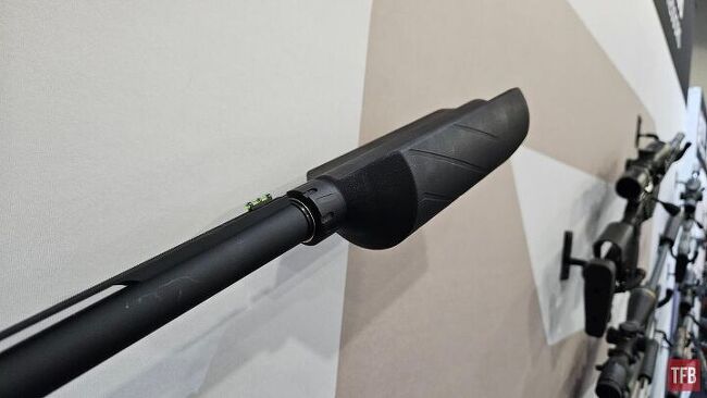 Close-up view of a rifle barrel fitted with a black muzzle device, displayed on a wall among other firearms.