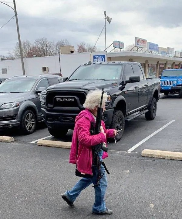 An elderly woman in a pink jacket walks through a parking lot carrying a rifle, with parked vehicles visible in the background.