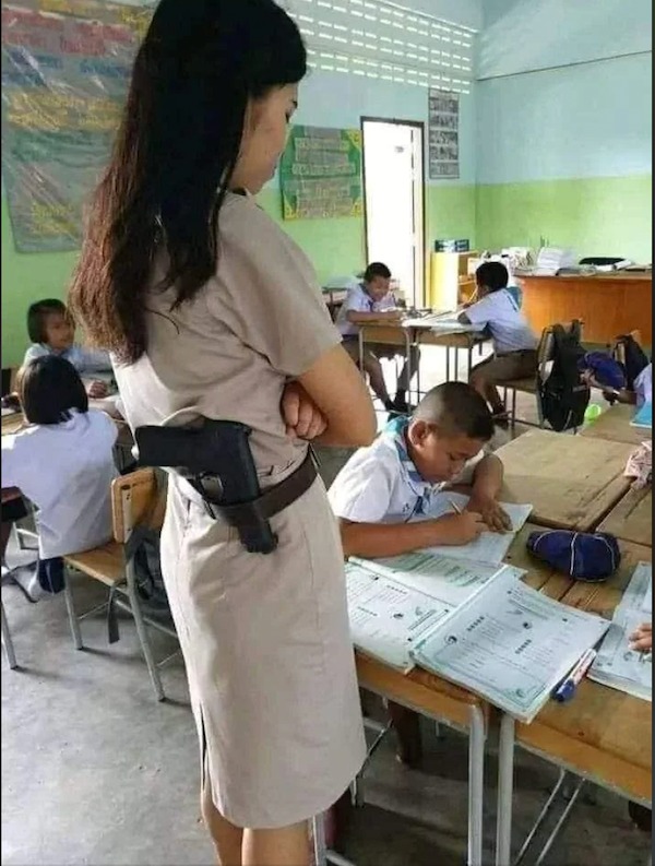 A teacher with a holstered gun on her belt stands by a student working at a desk in a classroom with other students.