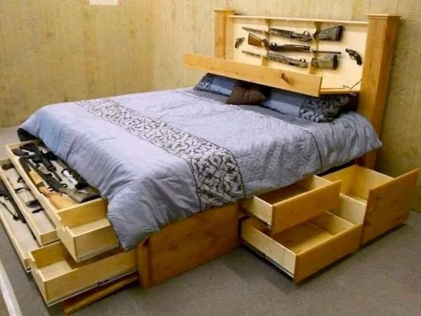 A wooden bed frame with multiple open drawers and compartments storing various guns and rifles, including hidden storage in the headboard and under the mattress.