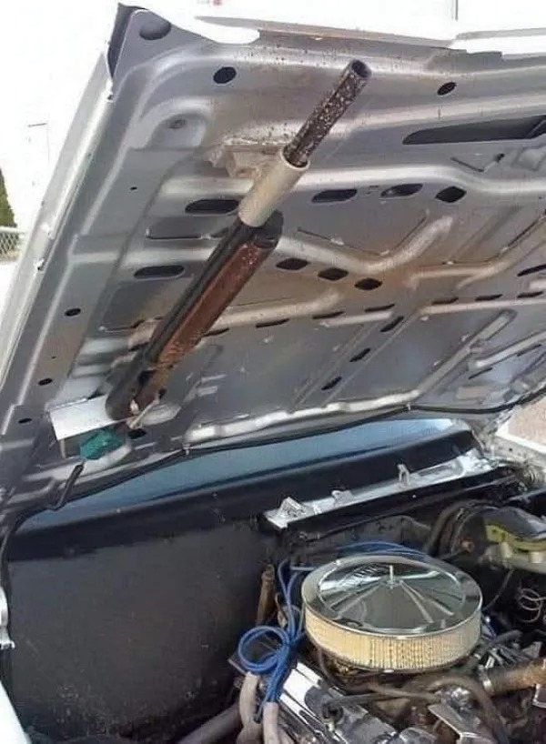 A car hood is propped open with a shotgun instead of a traditional hood support rod, showing the engine and wiring underneath.