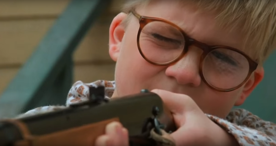 A young boy wearing glasses aims a BB gun with one eye closed, concentrating on his target.