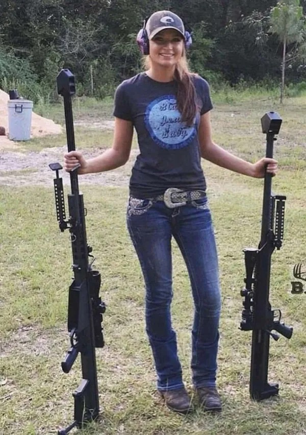 A woman standing outdoors on grass, smiling, and holding two large rifles, one in each hand, with trees and a trash bin visible in the background.