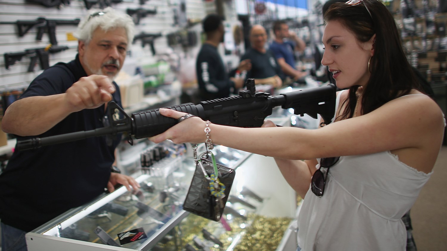 A woman holds and aims a rifle inside a gun shop as a man, possibly a store employee, gives instructions; other firearms and customers are visible in the background.