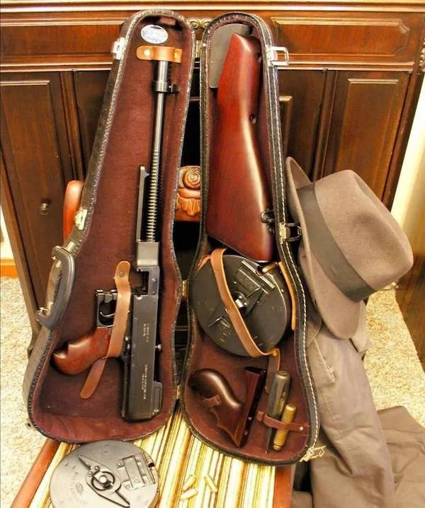 A violin case open to reveal a disassembled Thompson submachine gun, drum magazines, a brown fedora, and a gray coat beside it.