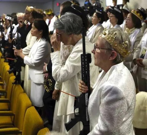 A group of women in white outfits and gold crowns stand in rows indoors, some holding rifles, during what appears to be a formal or ceremonial event.