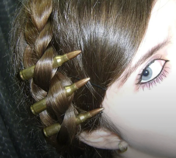A close-up of a woman's head showing a braid with rifle cartridges woven into her hair near the ear and eye.