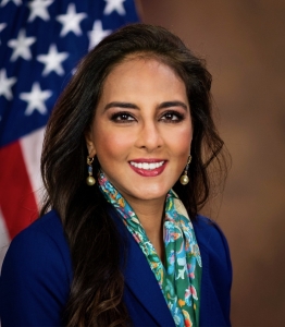 Harmeet K. Dhillon in a blue blazer and floral scarf smiles in front of a U.S. flag and a brown background.